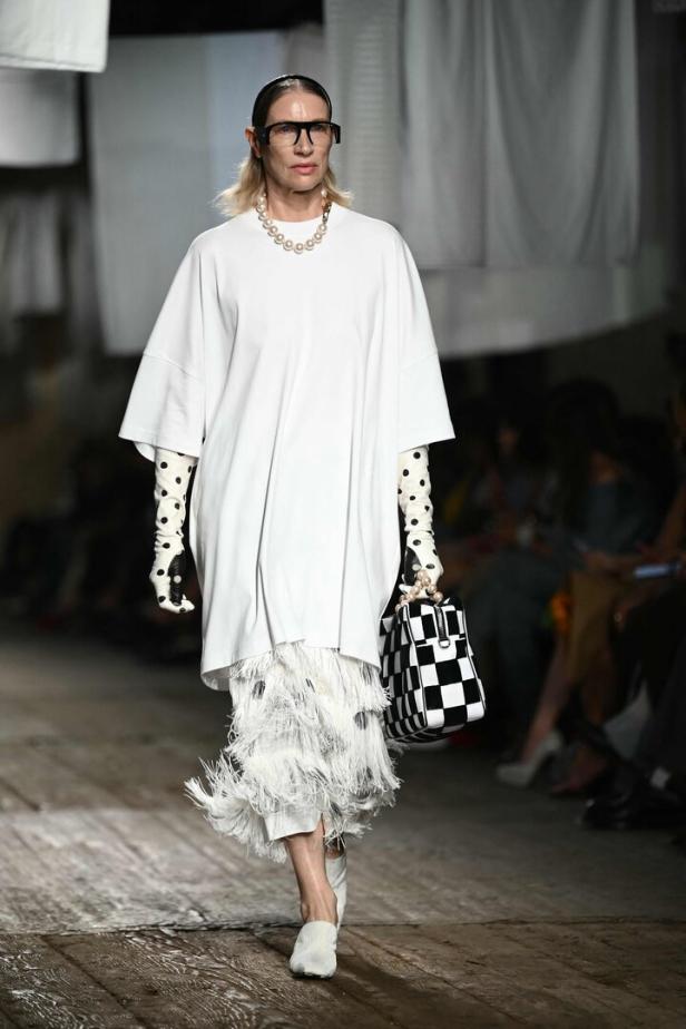 Eine Frau in einem weißen Kleid mit Fransen, gepunkteten Handschuhen und einer Schachbrettmuster-Tasche auf dem Laufsteg.