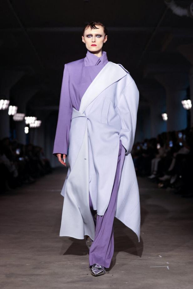 Ein Model präsentiert ein lila-blaues Outfit mit silbernen Schuhen auf einem Laufsteg.
