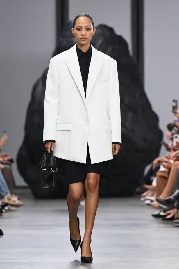 Eine Frau in einem weißen Blazer und schwarzen Kleid auf einem Laufsteg.