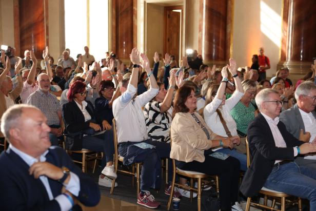 Viele Menschen sitzen in einem Saal und heben ihre Hände.