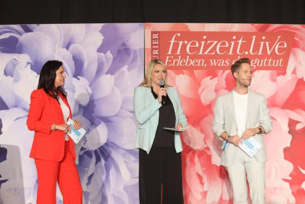 Drei Personen stehen auf einer Bühne vor einem Hintergrund mit Blumenmuster.