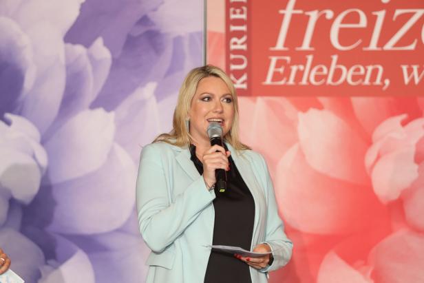 Eine blonde Frau spricht bei einer Veranstaltung in ein Mikrofon.