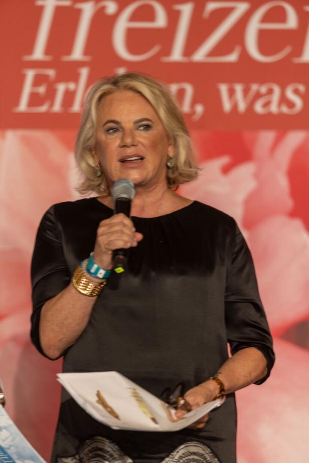 Eine Frau mit blonden Haaren spricht in ein Mikrofon und hält Papiere in der Hand.