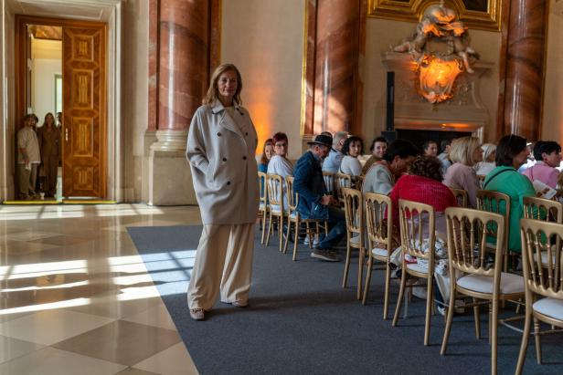 Eine Frau in einem hellen Mantel steht vor einer Gruppe sitzender Personen in einem prunkvollen Saal.