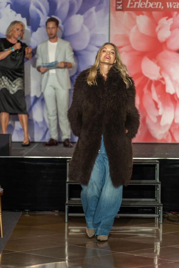 Eine Frau in einem braunen Pelzmantel und Jeans auf einer Bühne.