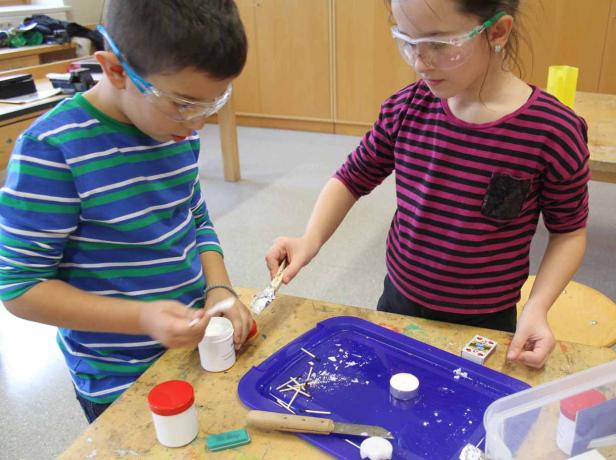 Zwei Kinder mit Schutzbrillen experimentieren mit Materialien auf einem Tisch.
