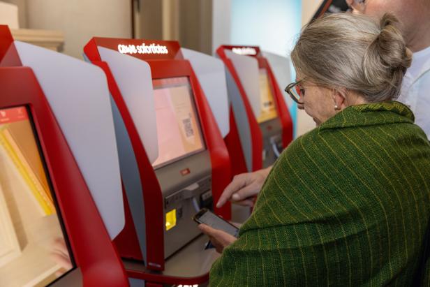 Eine ältere Frau benutzt einen Fotoautomaten und hält ein Smartphone in der Hand.