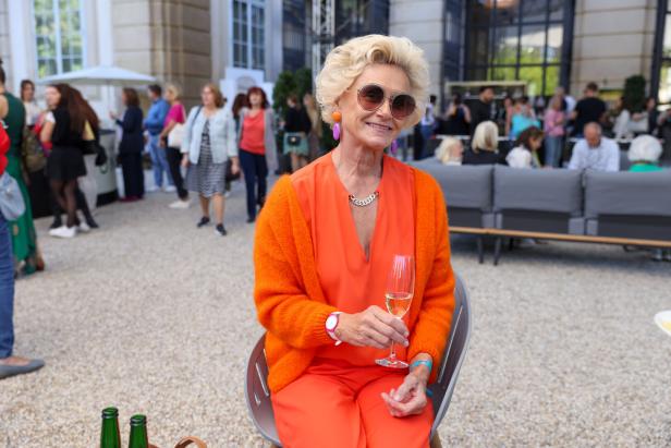 Eine ältere Frau in orangefarbener Kleidung sitzt mit einem Glas Sekt auf einer Veranstaltung.