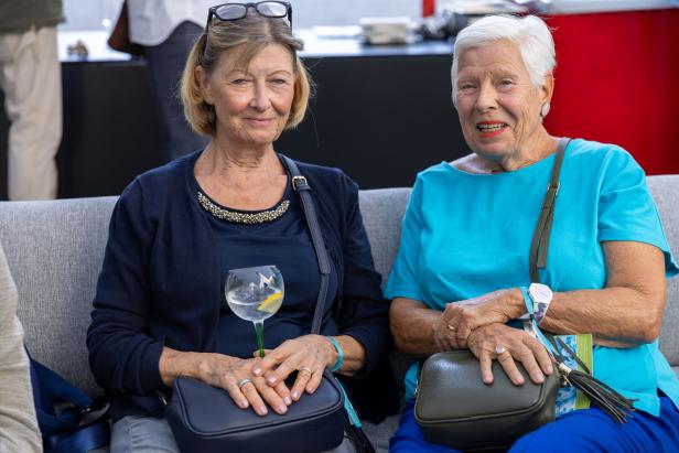 Zwei ältere Frauen sitzen auf einem Sofa, eine hält ein Getränk in der Hand.