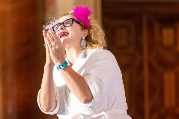 Eine Frau mit Brille, pinkfarbenem Kopfschmuck und Bademantel blickt erwartungsvoll nach oben.