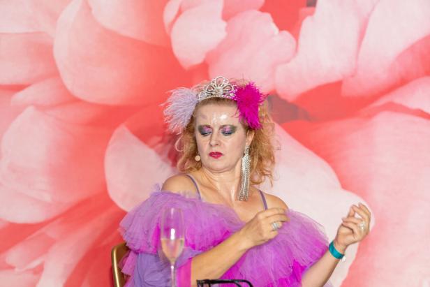 Eine Frau mit Tiara, Federn und üppigem, lila Tüll vor einem Blumenhintergrund.
