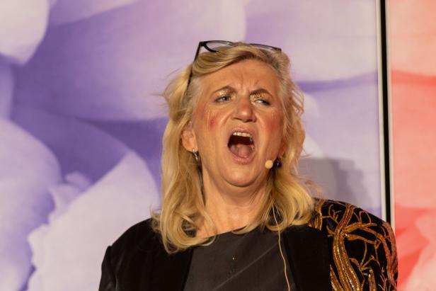 Eine Frau mit blonden Haaren und einem schwarzen Blazer mit goldenen Verzierungen schreit in ein Mikrofon.