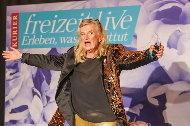Eine Frau mit blonden Haaren gestikuliert auf einer Bühne vor einem Werbebanner.