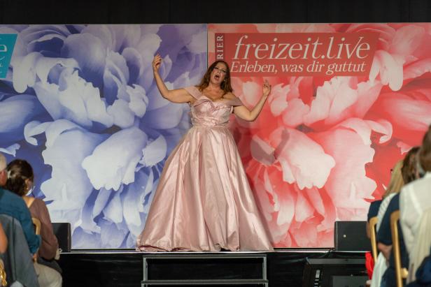 Eine Frau in einem rosa Kleid singt auf einer Bühne vor Publikum.