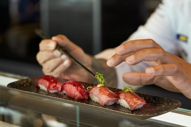 Ein Koch garniert Nigiri-Sushi mit grünen Zwiebeln auf einem länglichen Teller.