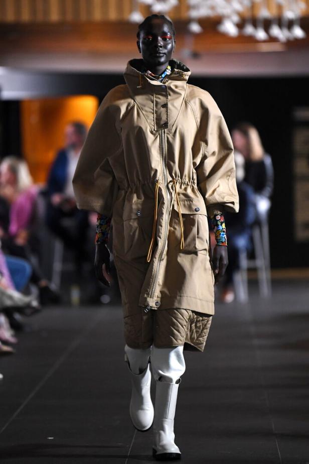 Eine dunkelhäutige Frau präsentiert einen beigen Parka und weiße Stiefel auf einem Laufsteg.