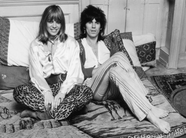 Anita Pallenberg und Keith Richards sitzen auf Decken und Polstern am Boden. Sie hat eine Leopardenhose und ein luftiges Hemd an, er eine lockere Leinenhose, ein Leinenhemd und eine Weste.