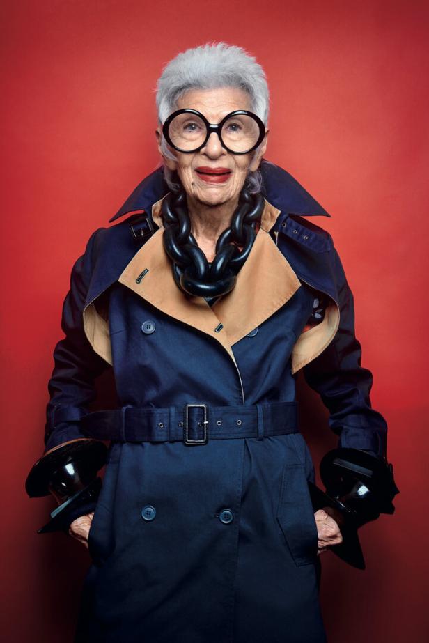 Iris Apfel mit übergroßer Brille, Trenchcoat und auffälliger Halskette vor rotem Hintergrund.