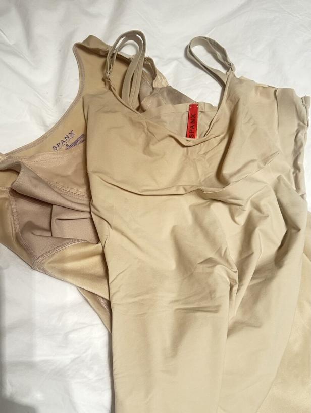 Zwei beigefarbene Shapewear-Oberteile der Marke Spanx liegen auf einem weißen Stoff.