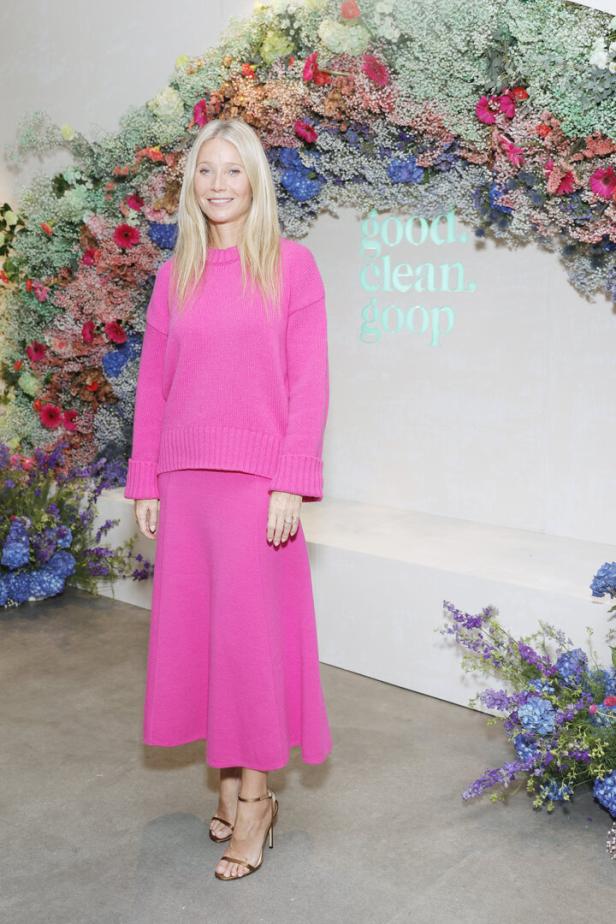 Gwyneth Paltrow in einem pinkfarbenen Ensemble vor einer Wand mit Blumen und dem Goop-Logo.