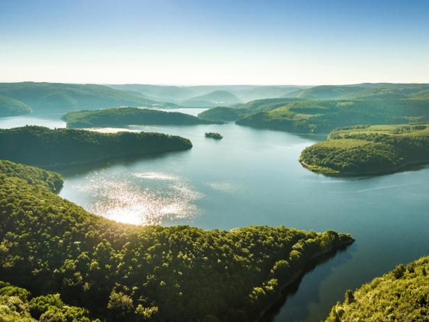 Luftaufnahme eines Sees mit bewaldeten Hügeln im Hintergrund.