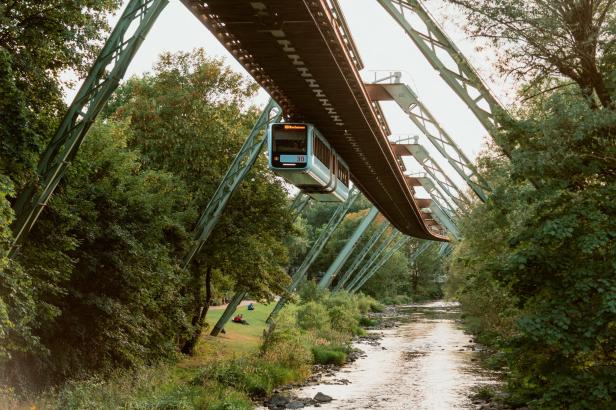 Die Wuppertaler Schwebebahn fährt über die Wupper, umgeben von Bäumen.