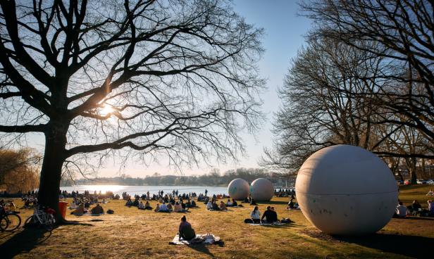 Menschen sitzen auf einer Wiese am Ufer eines Sees, im Hintergrund große Kugeln.