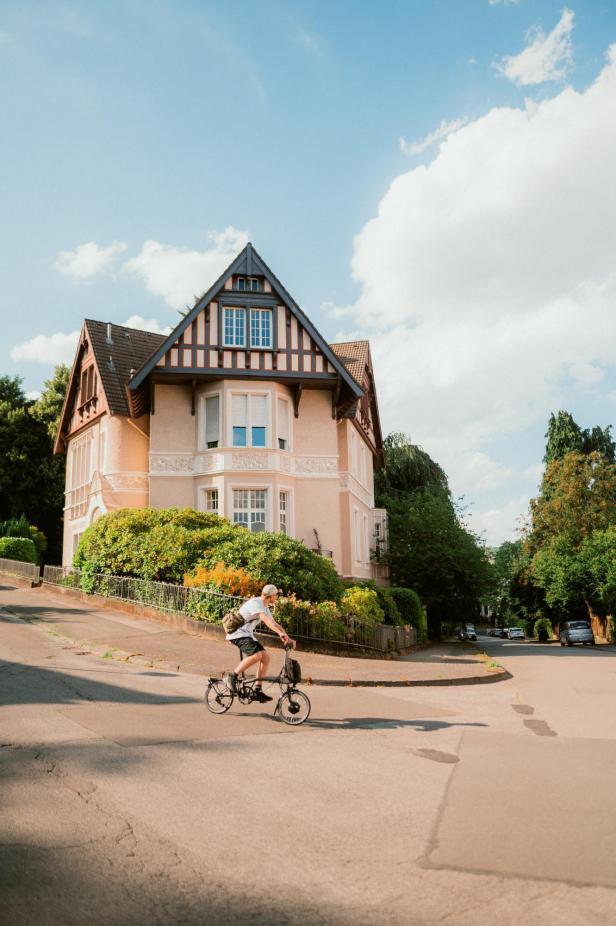 Ein Mann fährt mit einem Faltrad eine Straße entlang, im Hintergrund ein großes Haus.