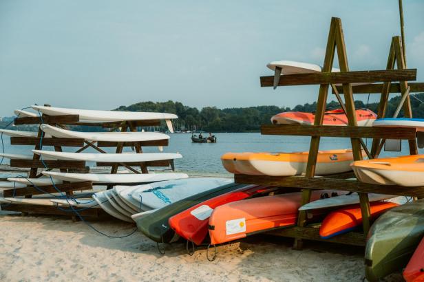 Mehrere Surfbretter und Kajaks liegen am Strand auf Holzgestellen.