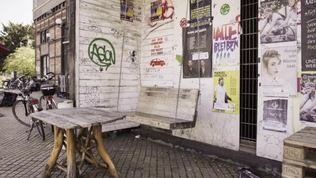 Eine hölzerne Schaukel hängt vor einer Wand mit Graffiti und Veranstaltungsplakaten.