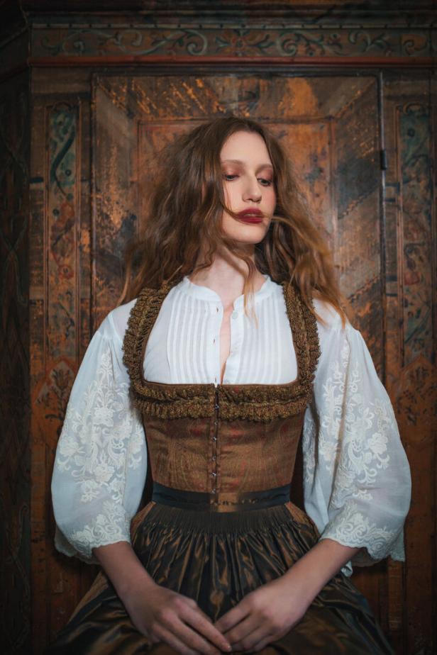 Eine junge Frau in Tracht steht vor einem verzierten Holzschrank.