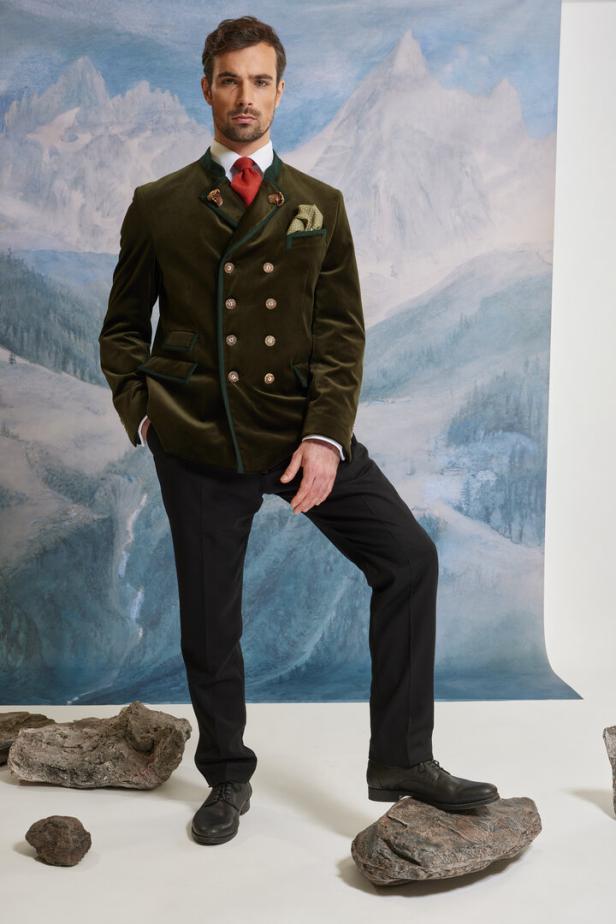 Ein Mann posiert in Trachtenjacke und schwarzer Hose vor einem Bergpanorama.