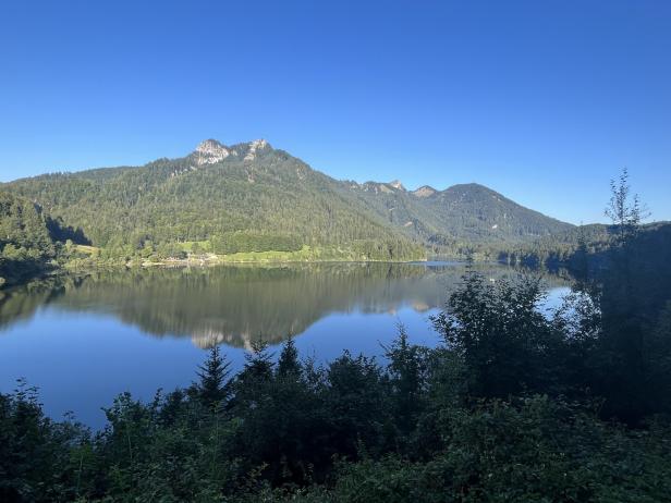 Der Thumsee liegt ruhig vor bewaldeten Bergen unter blauem Himmel.