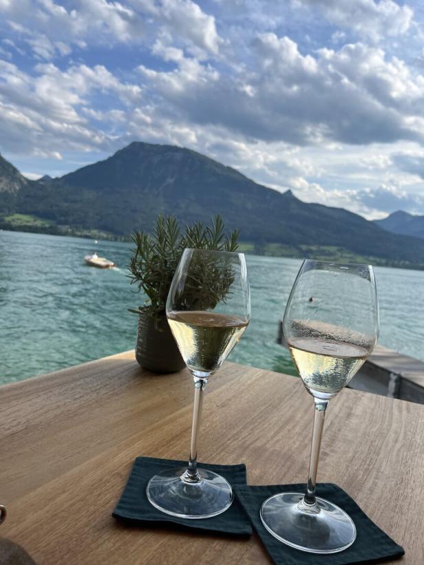 Zwei Gläser Weißwein stehen auf einem Tisch mit Blick auf einen See und Berge.