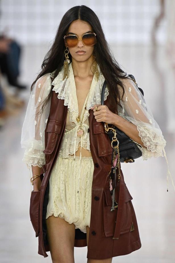 Eine Frau mit Sonnenbrille und braunem Ledermantel präsentiert ein Outfit mit Spitze.