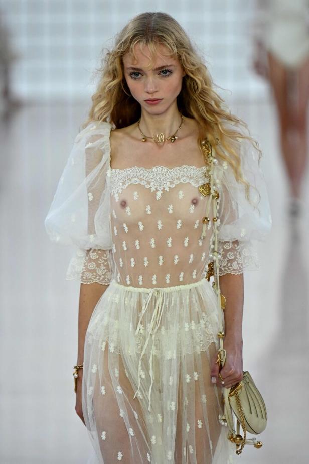 Ein Model mit blonden Haaren trägt ein transparentes, weißes Kleid mit Blumenmuster auf dem Laufsteg.
