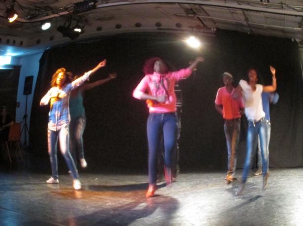 Eine Gruppe junger Leute tanzt auf einer Bühne vor einem dunklen Hintergrund.