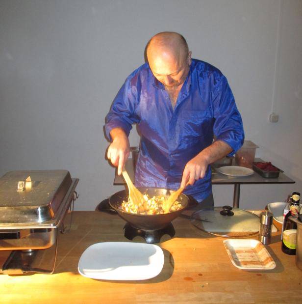Ein Mann mit blauem Hemd kocht mit zwei Holzlöffeln in einem Wok.