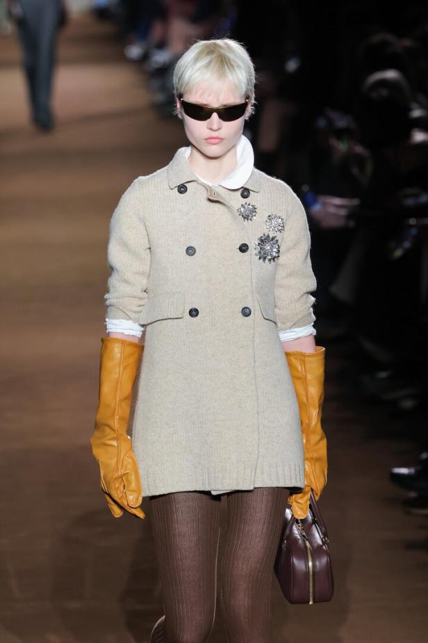 Ein Model mit blonden Haaren trägt einen beigen Mantel, braune Strumpfhosen und lange gelbe Handschuhe.
