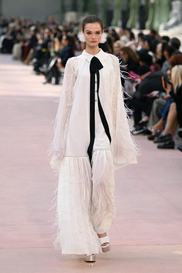 Ein Model präsentiert ein weißes Kleid mit Federn und einer schwarzen Schleife auf einem Laufsteg.