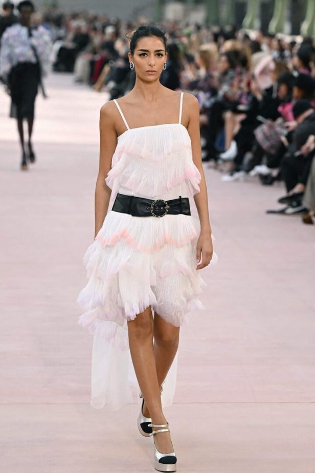 Ein Model präsentiert ein weißes, gestuftes Kleid mit schwarzem Gürtel auf dem Laufsteg.
