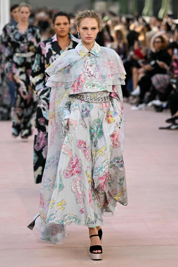 Ein Model auf dem Laufsteg trägt ein hellblaues, gemustertes Kleid mit passendem Cape.