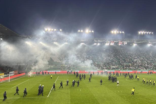 Polizeibeamte betreten ein Fußballfeld, das mit Rauch gefüllt ist, während Fans auf den Tribünen sitzen.