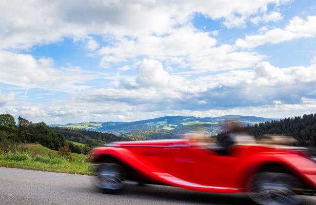Ein roter Oldtimer fährt auf einer Landstraße vor einer hügeligen Landschaft.