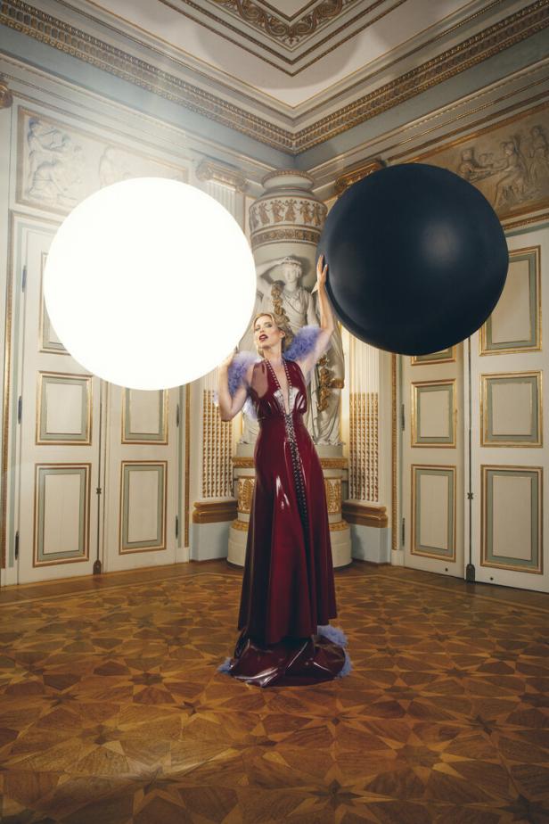 Kiera Chaplin in einem roten Kleid mit zwei großen Kugeln in einem verzierten Raum.