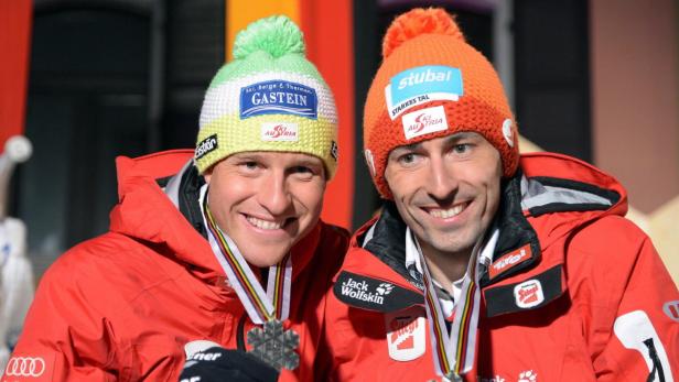 Zwei Skirennläufer mit Medaillen und Mützen mit Sponsorenlogos lächeln in die Kamera.