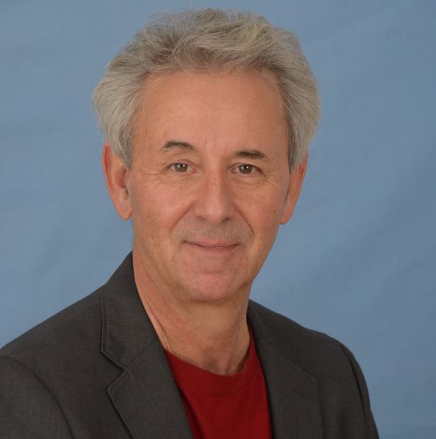 Porträt eines Mannes mit grauem Haar, der ein rotes Hemd unter einem grauen Sakko trägt.