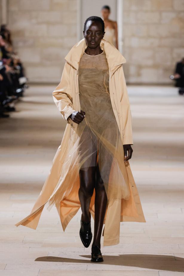 Eine dunkelhäutige Frau in einem beigen Mantel und einem transparenten Kleid auf einem Laufsteg.