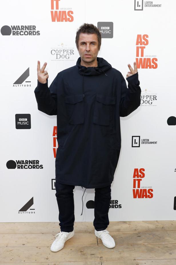 Liam Gallagher posiert vor einer Wand mit Logos und dem Titel „As It Was“.