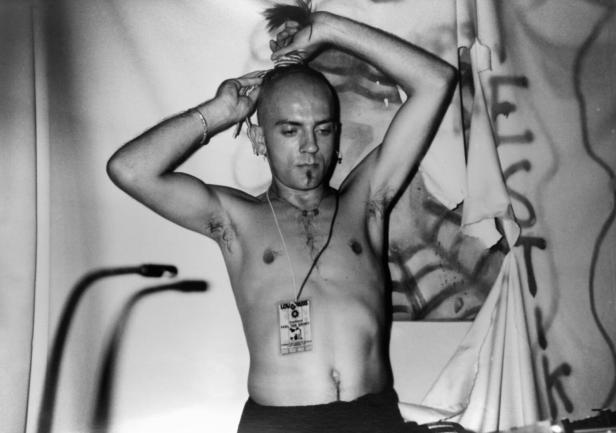 Der DJ Sven Väth mit nacktem Oberkörper und buddhistischer Mönchsfrisur vor einer besprühten Stoffwand.